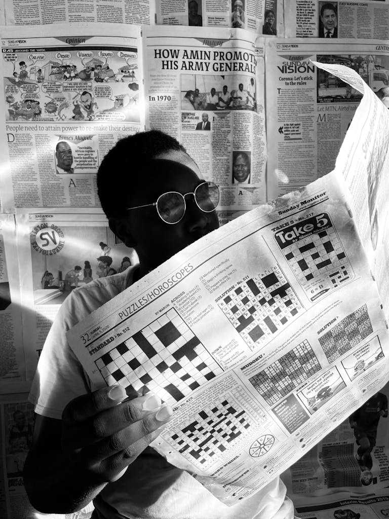 A person wearing glasses reads a crossword puzzle in a newspaper against a backdrop of various articles.