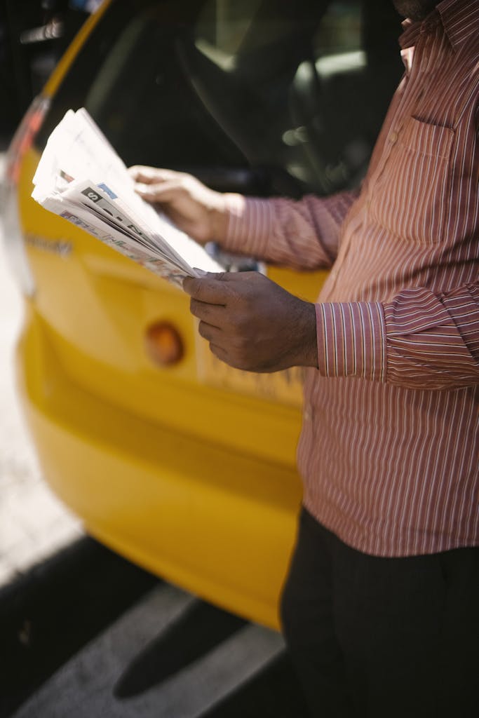 USA News TV A man in a striped shirt reads a newspaper beside a bright yellow taxi on a city street.