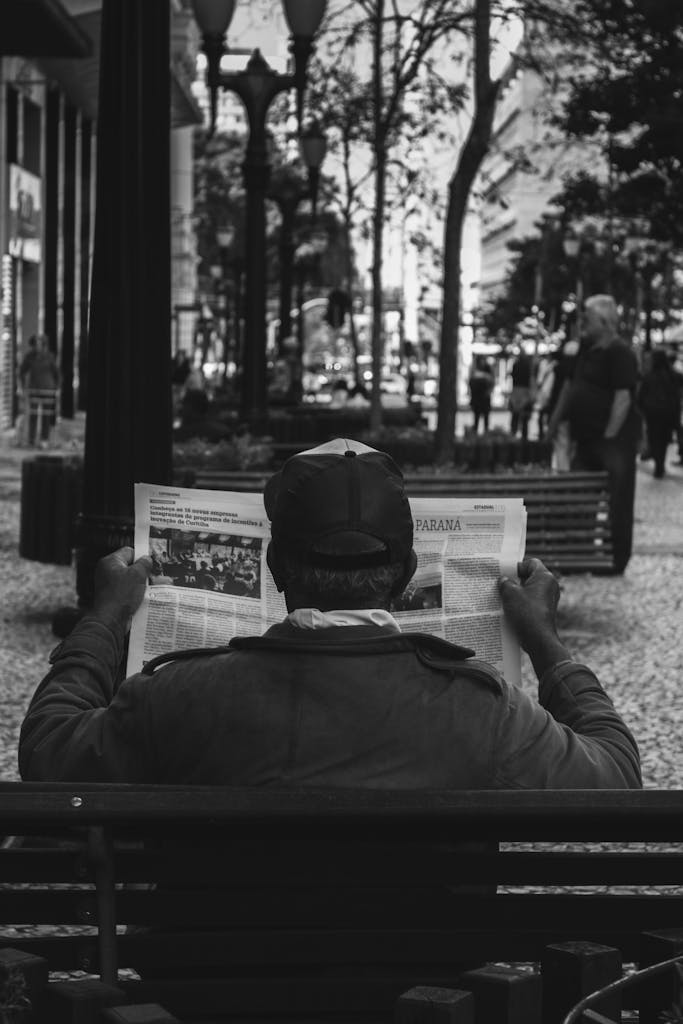 USA Today News A man sits on a bench in an urban park, reading a newspaper, captured in a black and white photo.