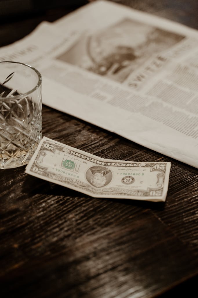 USA News Channel Live A vintage dollar bill next to a crystal glass on a wooden table and newspaper.