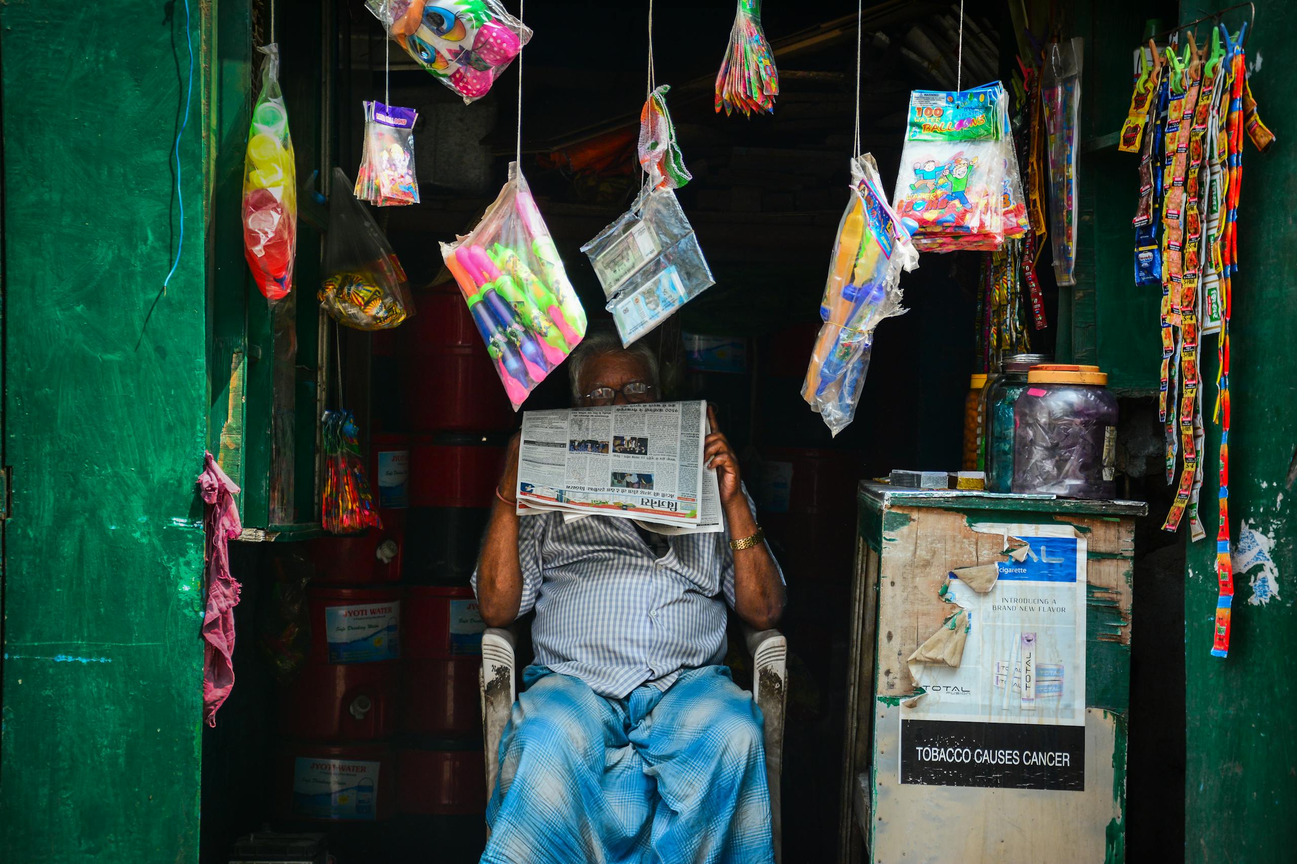 Gooding Idaho News An elderly man reading a newspaper in a vibrant market shop filled with hanging goods.