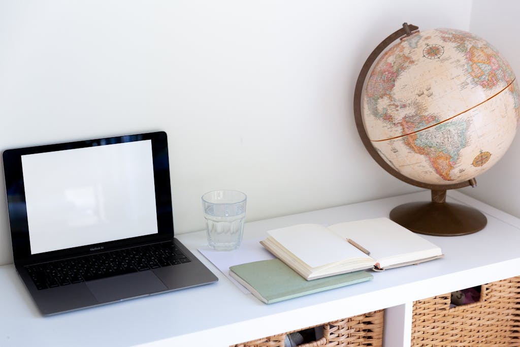 Globe and Mail Online Clean and organized workspace with a laptop, globe, notebooks, and glass of water on a white desk.