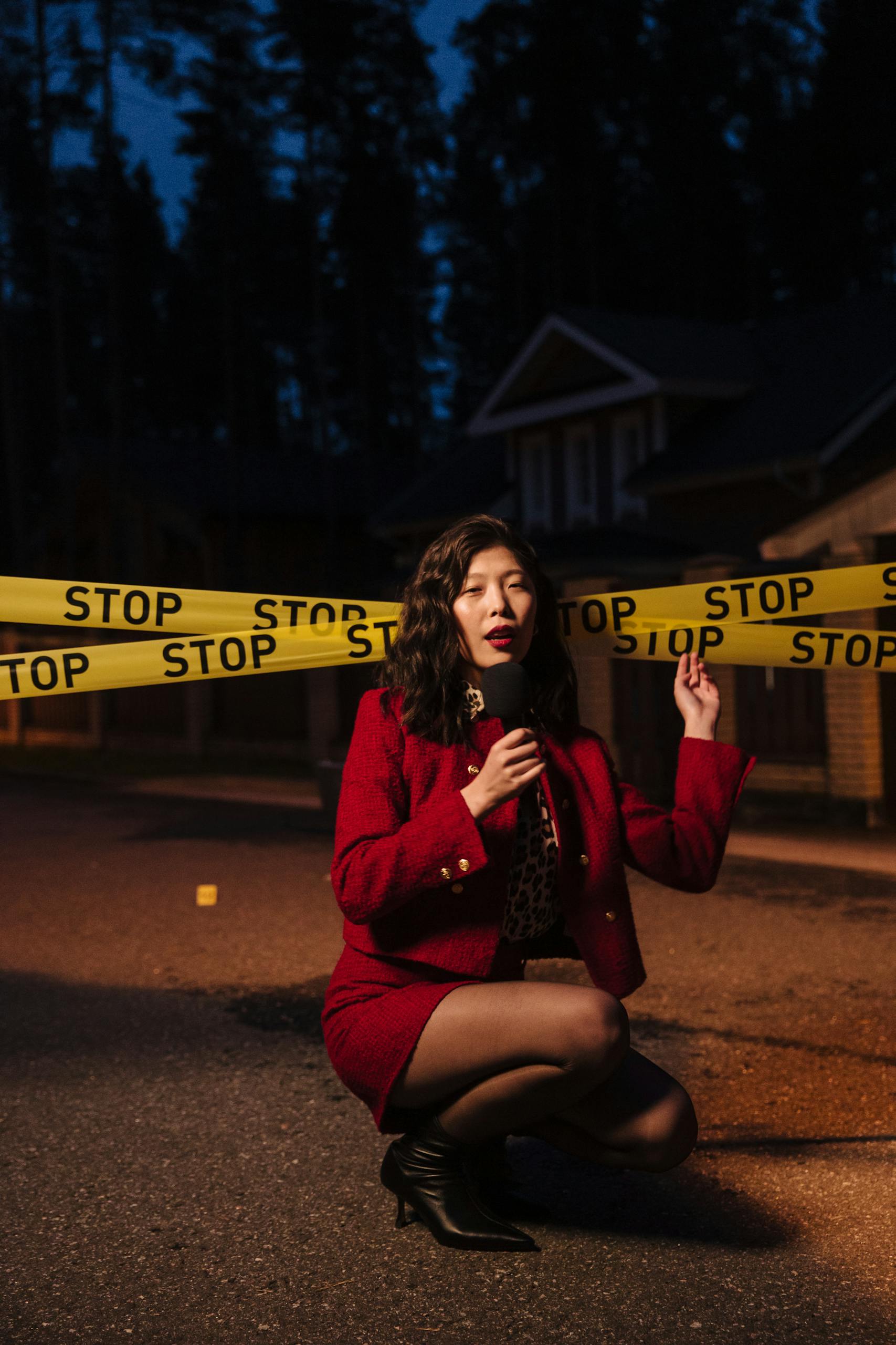 SosoActive News Female reporter at a crime scene with stop tape, crouching and holding a microphone at night.