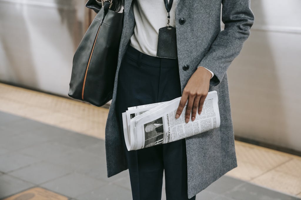From above of crop anonymous female employee in elegant outfit and name tag standing on platform of metro station with newspaper in hand