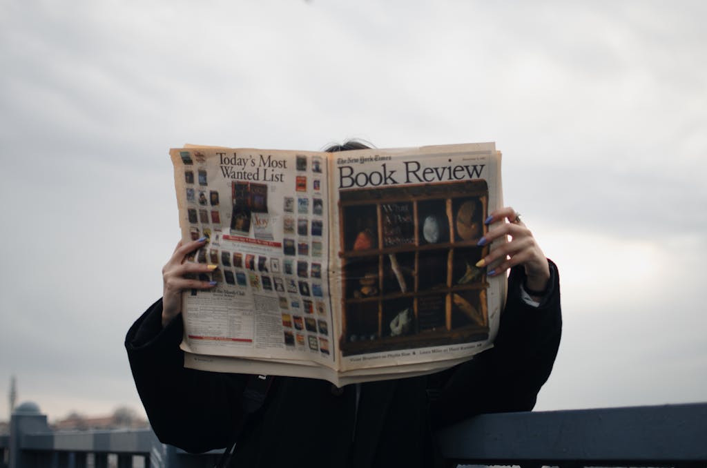 CBS News Individual holding a book review newspaper with colorful nails against a cloudy sky backdrop.