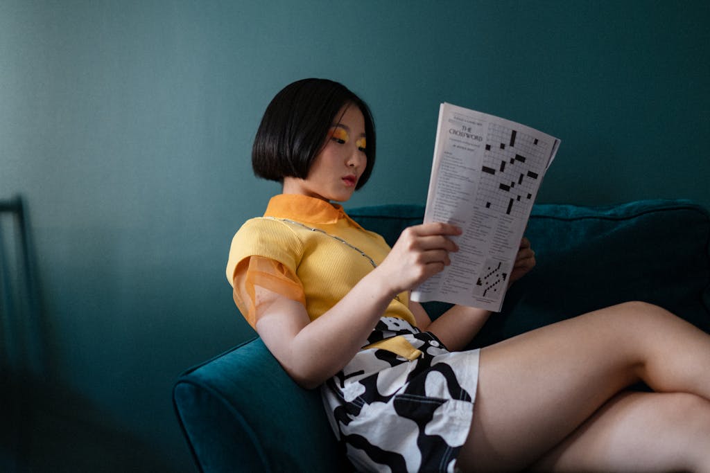 Stylish woman reading a crossword puzzle on a teal couch in a modern, relaxed setting.