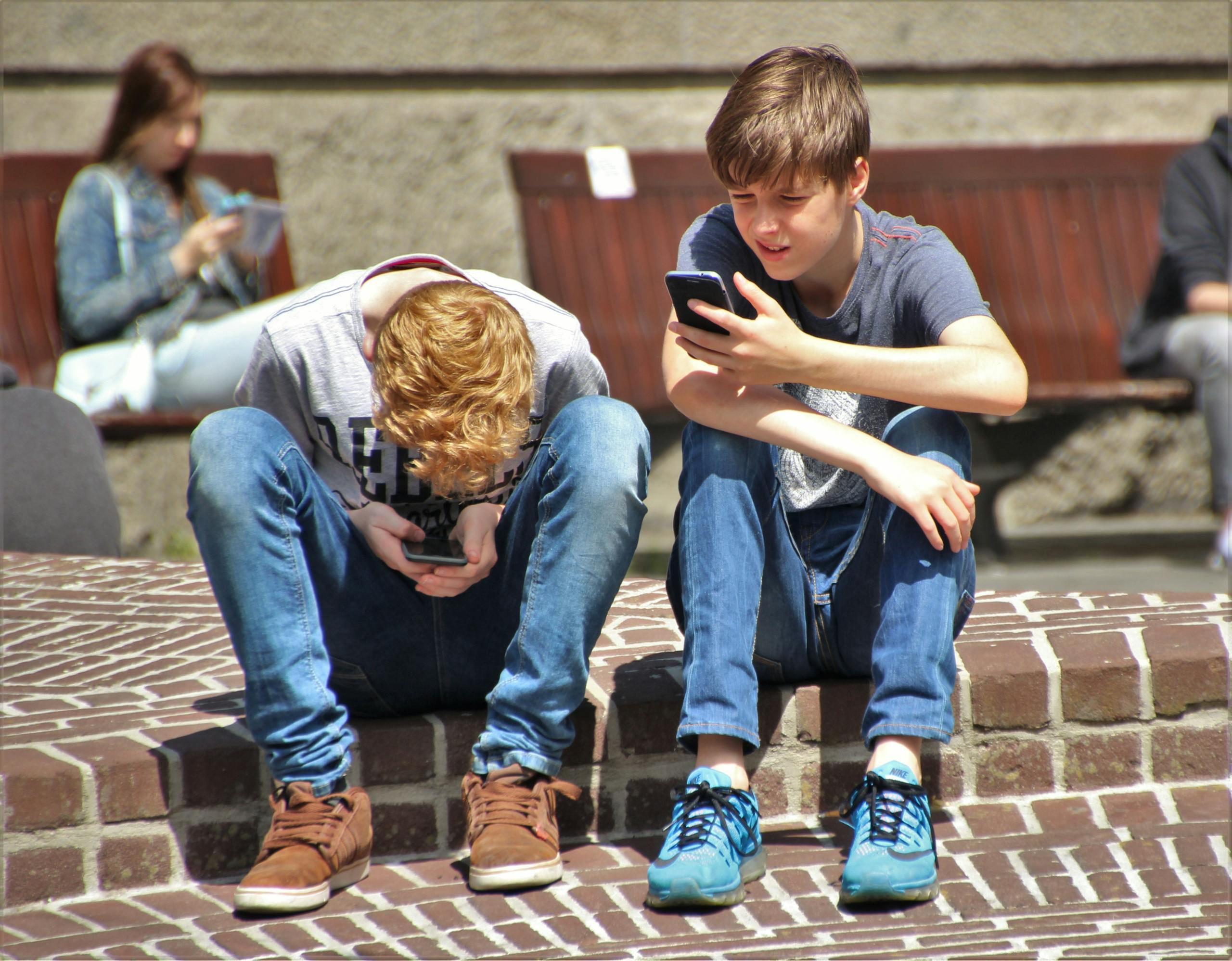 Newswell Two teenagers seated outdoors, focused on their smartphones, embracing modern technology.