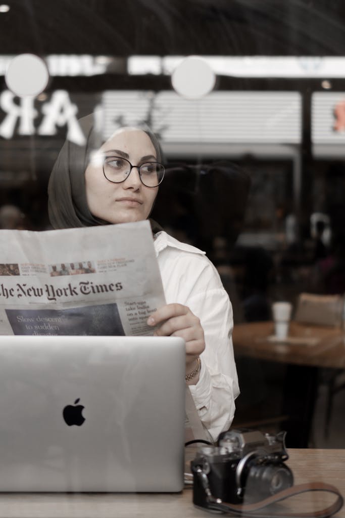 Woman in hijab reads a newspaper in a cafe, with a laptop and camera on the table.