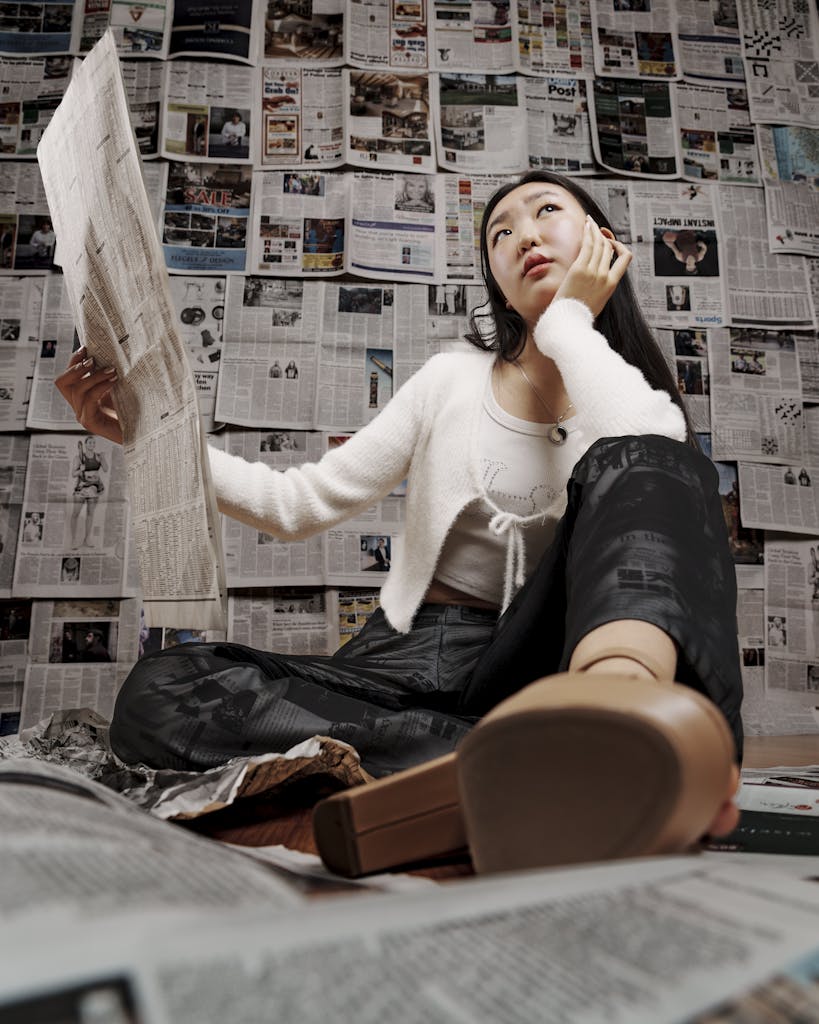 Young woman sitting and thoughtfully reading a newspaper indoors, surrounded by news articles.
