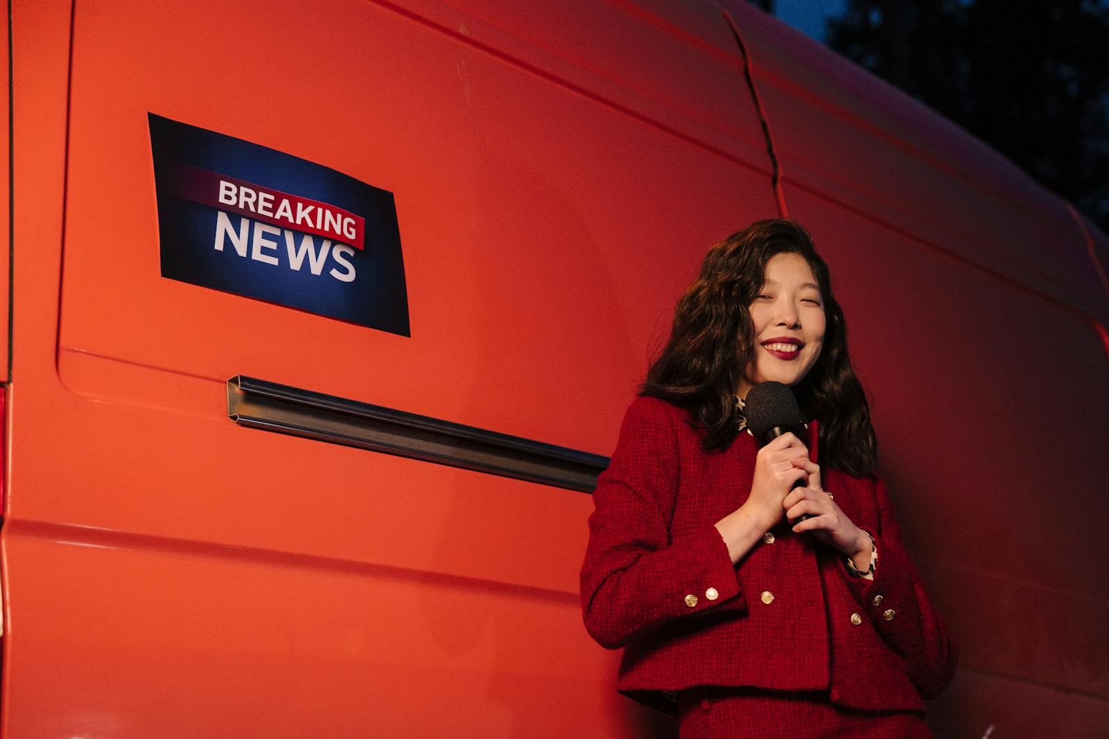 National Breaking NewsAsian female reporter smiling with microphone besides a news van at night.