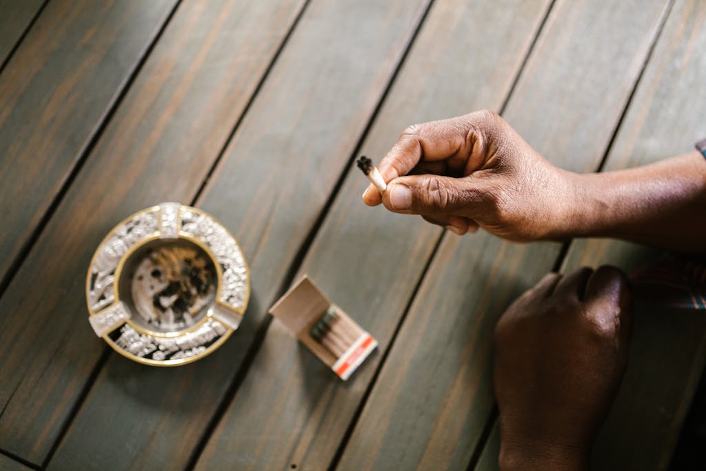 Movirz: A close-up shot of a person holding a marijuana joint above a wooden table with ashtray and matches.