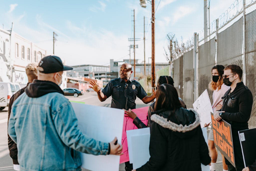 A police officer addresses a group of peaceful protesters holding signs on a city street under a clear sky.
