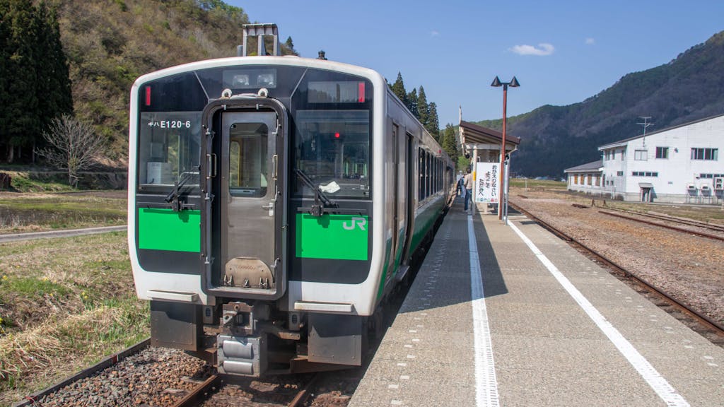 A train waits at a rural station surrounded by mountains and nature in Japan.