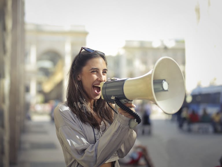 MSN News Cheerful young woman in a casual outfit shouting into a megaphone on a sunny day.