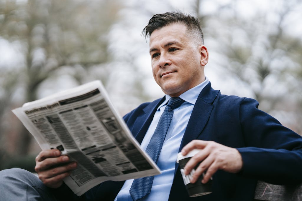 Stylish man in formal wear enjoys a drink while reading a newspaper outside.
