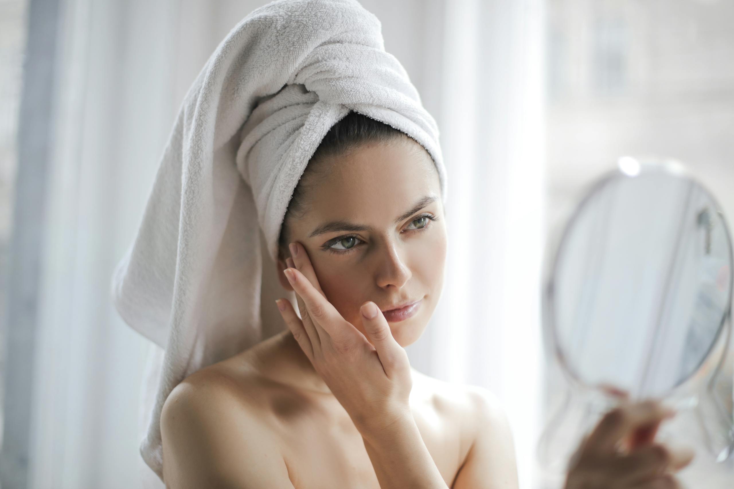 Well Health Tips in Hindi Wellhealthorganic Young female with bare shoulders and towel on head holding small mirror and checking skin with it in bathroom