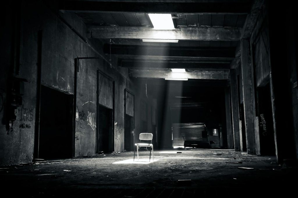 Sixmagazine  A haunting black and white photo of an abandoned warehouse with a solitary chair illuminated by sunlight through a window.