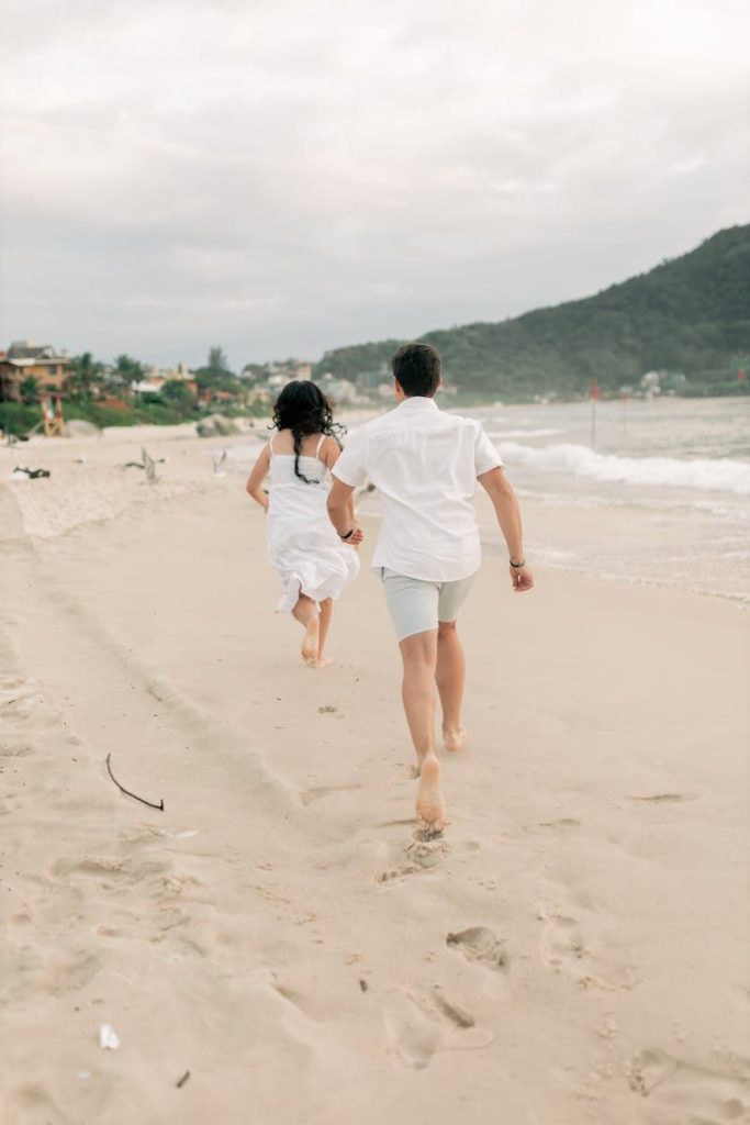 Couple in white clothing walking on a serene beach with picturesque views.