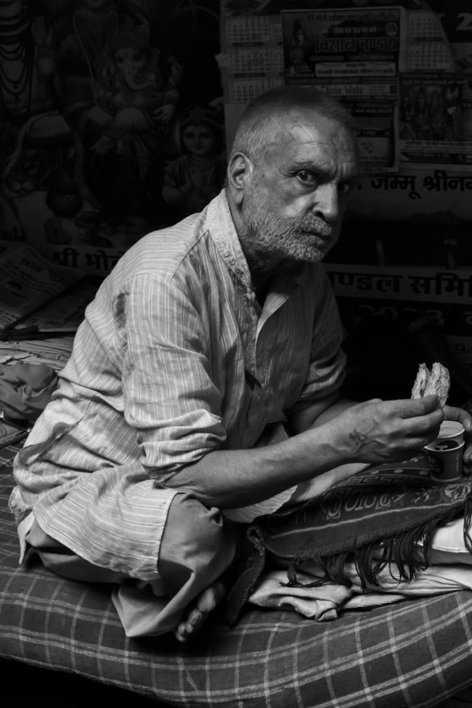 A contemplative elderly man eats a snack indoors in Delhi, India.