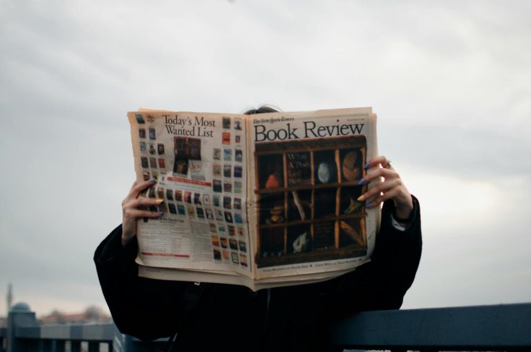 Rajkot Updates News Individual holding a book review newspaper with colorful nails against a cloudy sky backdrop.